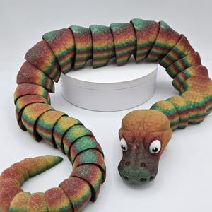 May include: A colourful, segmented snake toy with a green, brown, and orange head. The snake is made of a soft, textured material and has a segmented body.