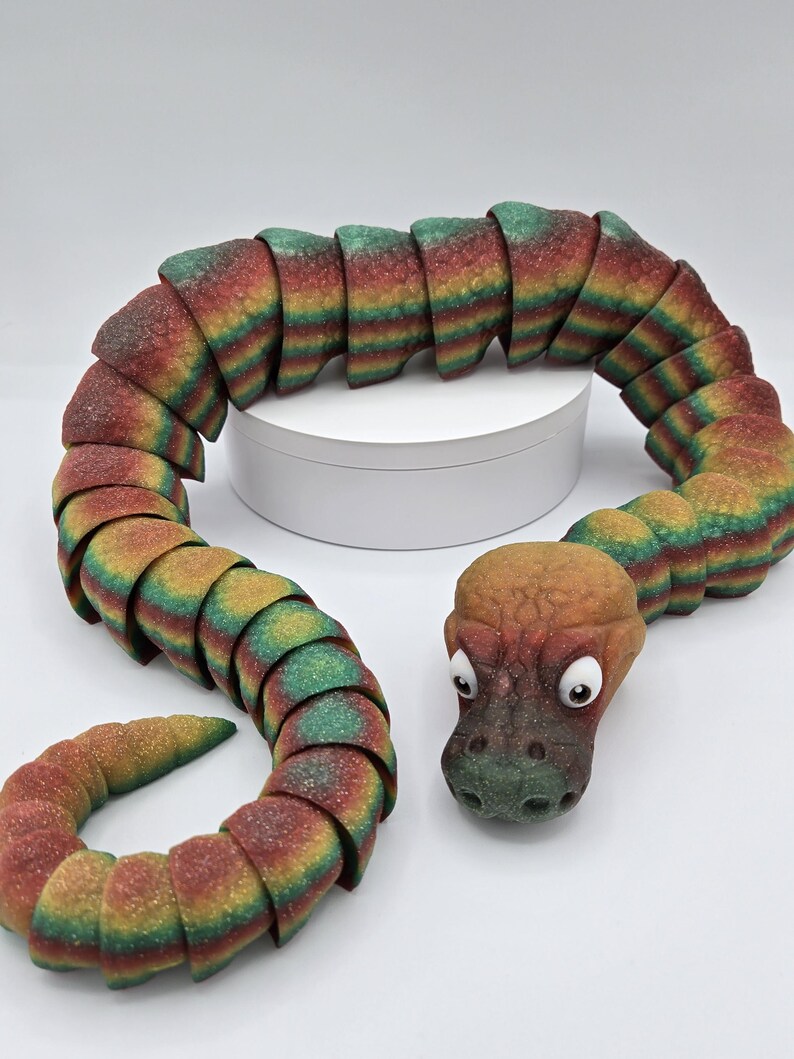 May include: A colourful, segmented snake toy with a green, brown, and orange head. The snake is made of a soft, textured material and has a segmented body.