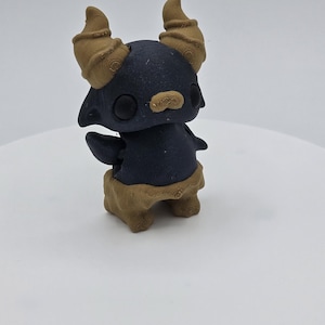 May include: A 3D-printed figurine of a stylized creature with a dark blue body and tan horns, eyebrows, and base. The figure has large, round eyes and small arms. The base has a textured, organic appearance.