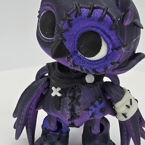 May include: A 3D-printed owl figurine in shades of purple and black. The owl has large, detailed eyes, stitched patches, and a white accent on its wrist. The design features a unique, textured appearance, with visible layer lines from the printing process.