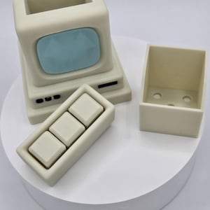 May include: A cream-colored, retro-style computer set, including a monitor with a blue screen, a keyboard with three square keys, and a small box. The set is displayed on a white, circular platform.