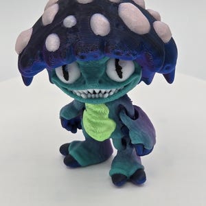 May include: A 3D-printed figurine of a mushroom-headed creature. The figure has a dark blue and pink spotted cap, a teal face with large white eyes, and a wide, toothy grin. Its body is teal and purple, with a green tie-like detail.
