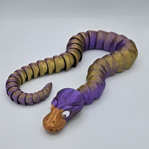 Może przedstawiać: Zabawka w kształcie węża, segmentowana, w kolorze fioletowym i złotym, z fakturowaną powierzchnią i dużymi, białymi oczami. Wąż jest zwinięty w spiralę.