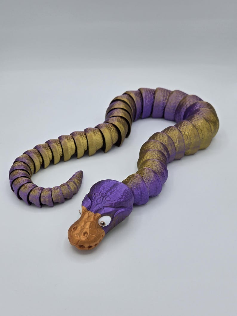 Może przedstawiać: Zabawka w kształcie węża, segmentowana, w kolorze fioletowym i złotym, z fakturowaną powierzchnią i dużymi, białymi oczami. Wąż jest zwinięty w spiralę.