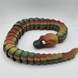 May include: A colourful segmented snake toy made of plastic. The snake is green, red, and yellow with a brown head. The head has a single white eye.