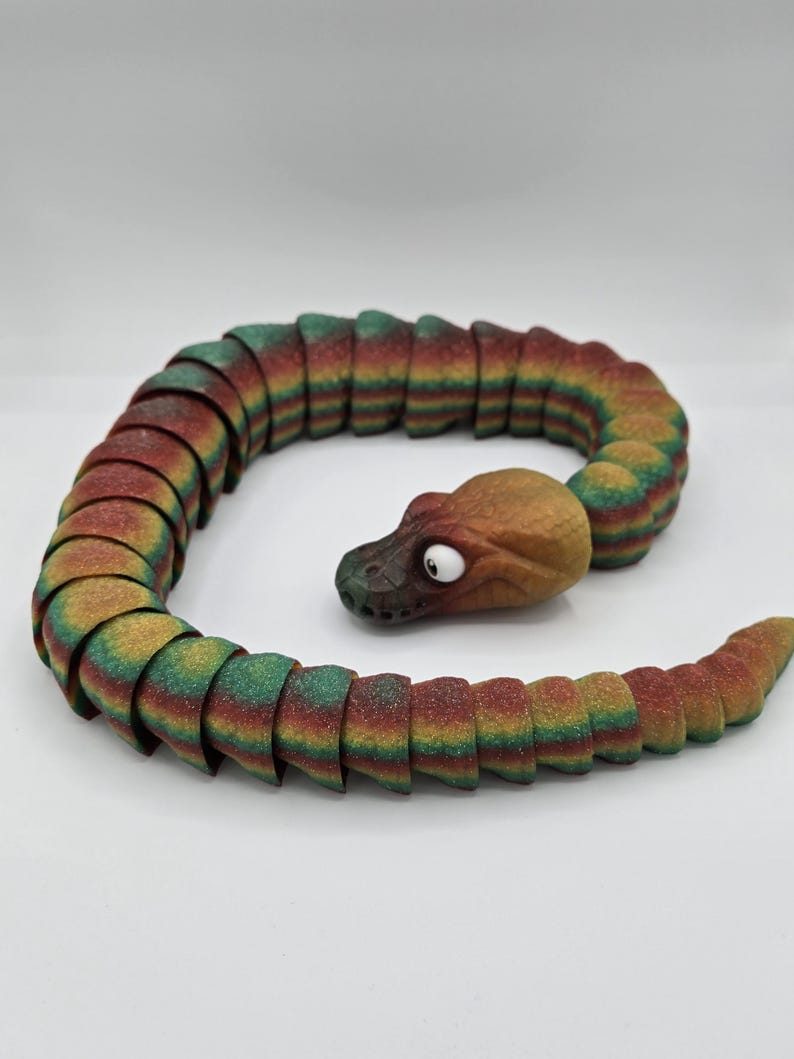 May include: A colourful segmented snake toy made of plastic. The snake is green, red, and yellow with a brown head. The head has a single white eye.