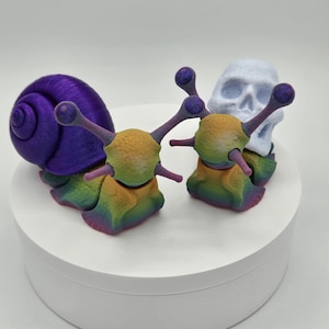May include: A whimsical sculpture featuring two snail-like creatures with rainbow-colored bodies and purple eye stalks. A purple snail shell and a light blue skull are also present. The sculpture is displayed on a white, circular platform.