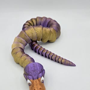 Może przedstawiać: Zabawka w kształcie węża, segmentowana, w kolorze fioletowym i złotym, z fakturowaną, pomarańczową głową przypominającą kaczkę. Zabawka jest wykonana z plastiku i ma segmentowane ciało.