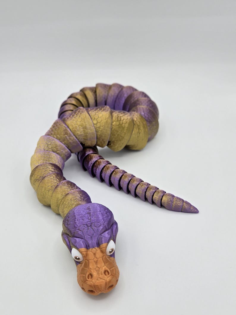 Może przedstawiać: Zabawka w kształcie węża, segmentowana, w kolorze fioletowym i złotym, z fakturowaną, pomarańczową głową przypominającą kaczkę. Zabawka jest wykonana z plastiku i ma segmentowane ciało.