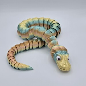 Może przedstawiać: Zabawka w kształcie węża, segmentowana, wielokolorowa, z ż&oacute;łtą głową i dużymi, okrągłymi oczami. Wąż wykonany jest z fakturowanego, elastycznego materiału i jest zwinięty w spiralę.