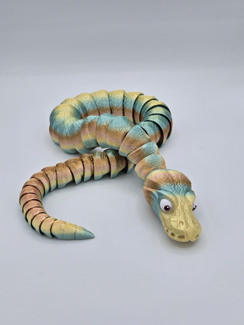 Może przedstawiać: Zabawka w kształcie węża, segmentowana, wielokolorowa, z ż&oacute;łtą głową i dużymi, okrągłymi oczami. Wąż wykonany jest z fakturowanego, elastycznego materiału i jest zwinięty w spiralę.