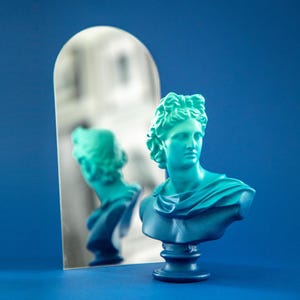 Könnte beinhalten: Eine blaue und grüne, verlaufende Büste eines Menschen mit einem klassischen griechischen Haarschnitt. Die Skulptur steht auf einem Sockel und vor einem Spiegel.