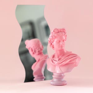 Könnte beinhalten: Eine rosafarbene Büste einer klassischen Figur steht vor einem wellenförmigen Spiegel, der die Skulptur reflektiert. Die Büste steht auf einem Sockel, vor einem sanften rosa Hintergrund. Die Skulptur hat detaillierte Gesichtszüge und drapierte Stoffe.