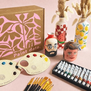 May include: A craft kit box labelled Paint date. with pink floral patterns, two wooden palettes with paint dabs, a set of paint tubes, several brushes, and four ceramic vases featuring hand-painted designs including a face, watermelon, and lemons.