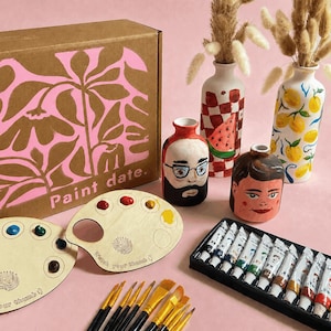 May include: A craft kit box labelled Paint date with a pink floral pattern, two wooden palettes with paint wells and the text Watch your thumb, two painted ceramic vases, two painted ceramic heads, and a set of paint tubes with brushes.