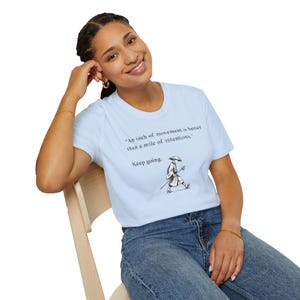 Peut inclure: Un t-shirt bleu clair à manches courtes avec le texte imprimé en noir "An inch of movement is better than a mile of intentions." et "Keep going." au-dessus d'une illustration en noir et blanc d'une personne portant un chapeau conique.