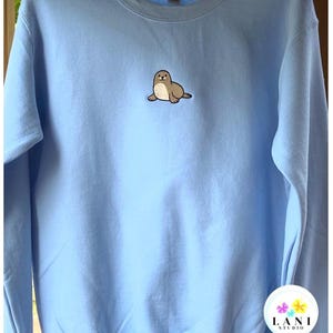 May include: A pale blue sweatshirt with a crew neck and long sleeves. It has a small embroidered seal in brown and cream on the front. The brand logo "LANI STUDIO" is visible in the bottom right corner.
