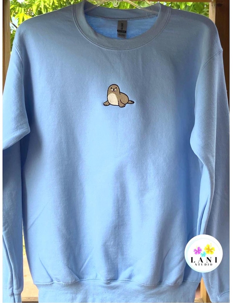 May include: A pale blue sweatshirt with a crew neck and long sleeves. It has a small embroidered seal in brown and cream on the front. The brand logo "LANI STUDIO" is visible in the bottom right corner.