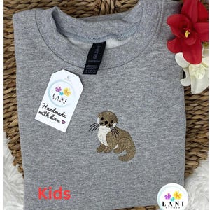 May include: A heather grey crewneck sweatshirt featuring an embroidered brown and white otter on the front. A white tag attached to the collar displays the text LANI STUDIO and Handmade with love. The word Kids is printed in red at the bottom.