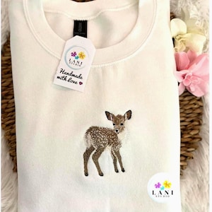 K&ouml;nnte beinhalten: Ein wei&szlig;es Sweatshirt mit Rundhalsausschnitt und einem gestickten Reh-Design. Das Reh ist braun und wei&szlig; mit dunkelbraunen Beinen und Ohren. Ein Etikett mit dem Text "Handmade with love" und dem Logo "LANI STUDIO" ist angebracht.