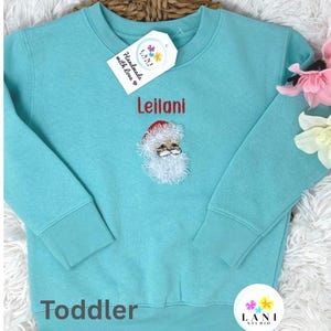 May include: A turquoise toddler sweatshirt featuring the name "Leilani" in red embroidery. It has a Santa Claus face design and a tag that says "Handmade with love". The sweatshirt has a crew neck and long sleeves.
