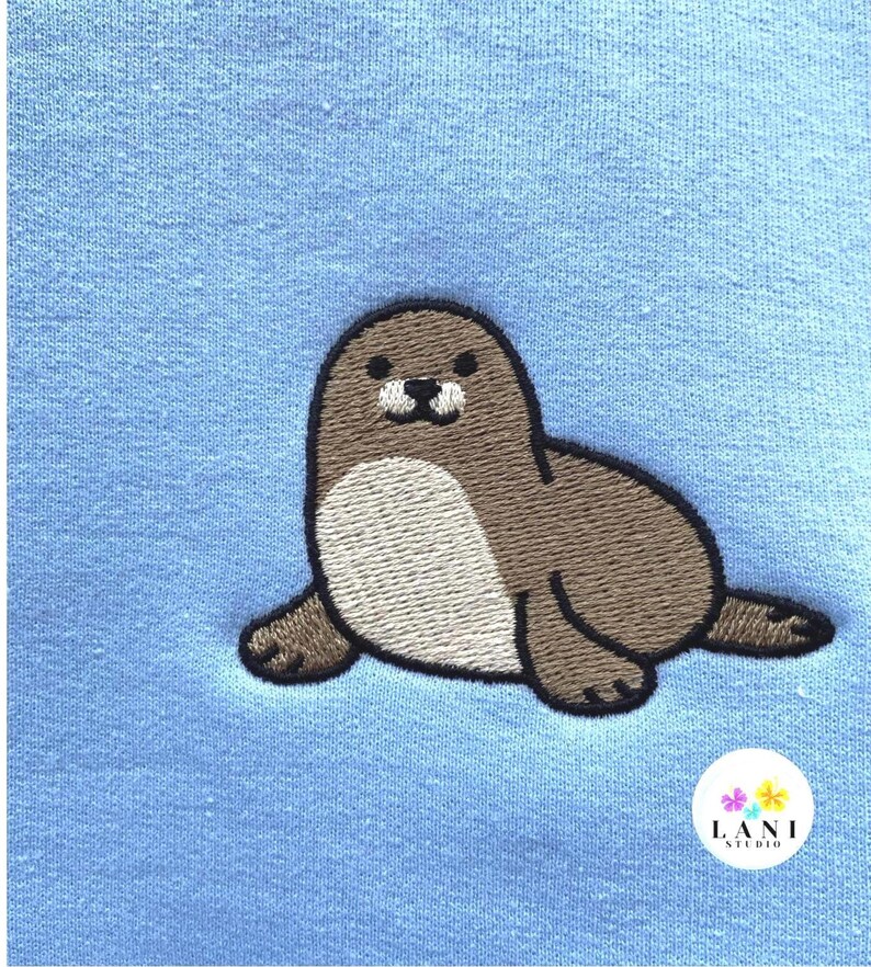 May include: Embroidered seal design in brown, tan, and black on a light blue fabric. The seal is lying down with its flippers extended. The design is from Lani Studio.