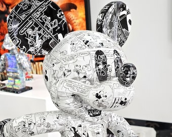 Op de afbeelding: Een wit Mickey Mouse beeldje bedekt met zwart-witte comicboekpanelen. Het beeldje is geposeerd met één arm uitgestrekt en de andere arm gebogen in de elleboog. Het beeldje is gemaakt van een glad, glanzend materiaal.