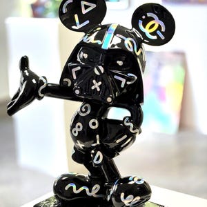Peut inclure: Sculpture noire représentant un personnage hybride de Dark Vador et d'une célèbre souris. La figurine a une finition brillante avec des accents irisés et est montée sur une base noire et blanche mouchetée. La figurine est en pose avec un bras tendu.