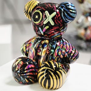 Puede incluir: Figura decorativa de oso con acabado brillante y diseño abstracto. El oso es predominantemente negro, adornado con patrones vibrantes de colores arcoíris, caras y rayas. Las orejas y el vientre tienen diseños únicos. La figura está sobre una superficie blanca.