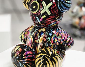 May include: A colorful, abstract art bear figurine with a glossy finish. The bear features a black base with vibrant patterns of rainbow colors, faces, and stripes. The bear's ears and belly have unique designs. The figurine is sitting on a white surface.