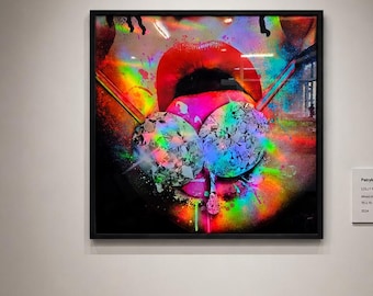 Op de afbeelding: Een vierkante ingelijste kunstdruk aan een witte galerijmuur, met een levendige, veelkleurige afbeelding van lippen met een regenboogspectrum en glitterachtige deeltjes, weergegeven naast een klein wit informatiebordje.