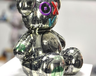 Op de afbeelding: Een zilverkleurige teddybeer sculptuur met dollarbiljet details. De beer heeft een paars oog, een rode X en blauwe en groene V-vormige accenten. De beer zit op een witte ondergrond.
