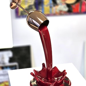 Puede incluir: Una escultura de una copa de vino de metal dorado, que parece verter líquido rojo en forma de salpicadura. La base es roja con siluetas negras. La obra de arte se exhibe sobre una superficie blanca.