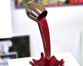 Op de afbeelding: Een sculptuur van een wijnglas van goudkleurig metaal, dat schijnbaar rode vloeistof in een spatvorm giet. De basis is rood met zwarte silhouetten. Het kunstwerk wordt op een wit oppervlak tentoongesteld.
