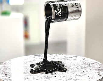 May include: A silver and black soup can sculpture suspended in mid-air, pouring a thick, glossy black liquid onto a circular base covered in a black and white comic book print. The can features the text Campbell's Condensed and Famous Soup.