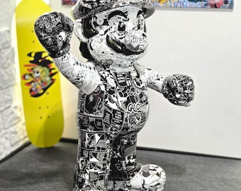 Op de afbeelding: Een zwart-wit pop-art sculptuur van een personage met een hoed met een "M", een overall en handschoenen. De figuur is bedekt met stripboekachtige graphics. Een gele skateboard met een afbeelding staat op de achtergrond.