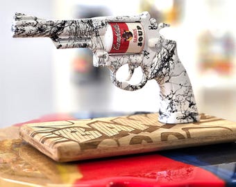 Op de afbeelding: Decoratieve handvuurwapensculptuur met een wit en zwart spetterpatroon. De cilinder is een Campbell's soepblik. De sculptuur rust op een houten voet met gegraveerde tekst. Een uniek kunstwerk.