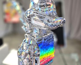 Op de afbeelding: Een metalen sculptuur van een stripfiguur, bedekt met stripverhalen. De sculptuur heeft een regenboogkleurig gedeelte en de tekst "Beep Beep" en "Flyweight Wonders". De sculptuur staat op een zwarte basis.
