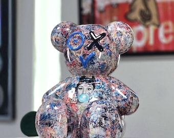May include: A colorful resin bear figurine with graffiti-style paint splatters in blue, red, and white. The bear has blue and black facial details, and a portrait of a woman blowing a bubble.