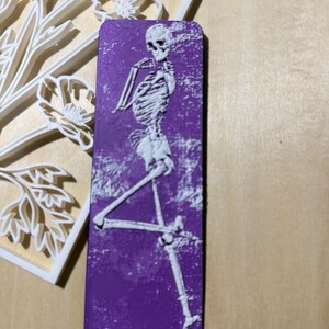 Puede incluir: Marcapáginas rectangular morado con un diseño de esqueleto blanco. El esqueleto está en una pose de baile. Un diseño floral blanco está en el fondo.
