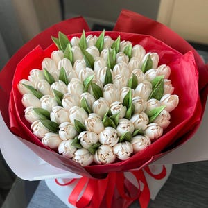 May include: A bouquet of white tulips with green leaves, wrapped in red and white paper, tied with a red ribbon. The flowers are tightly arranged, creating a full, lush appearance. The vibrant red paper contrasts with the delicate white tulips.