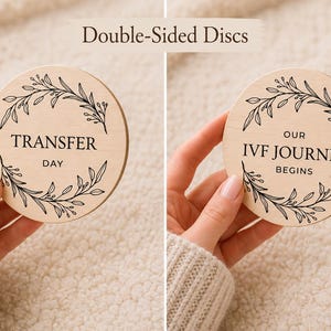 Pode incluir: Dois discos de madeira com texto preto e desenhos de folhas. Um disco diz "TRANSFER DAY", o outro diz "OUR IVF JOURNEY BEGINS". Os discos s&atilde;o segurados por m&atilde;os, com um fundo bege.