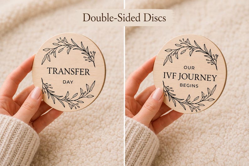 Pode incluir: Dois discos de madeira com texto preto e desenhos de folhas. Um disco diz "TRANSFER DAY", o outro diz "OUR IVF JOURNEY BEGINS". Os discos s&atilde;o segurados por m&atilde;os, com um fundo bege.