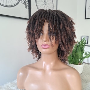 May include: A brown wig with a curly, loc-style texture is displayed on a mannequin bust. The wig has varying shades of brown and is styled with a side part. The mannequin has a neutral skin tone and is set against a white background.