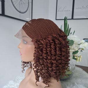 Peut inclure: Perruque tressée marron avec des extrémités bouclées sur une tête de mannequin. La perruque a un devant en dentelle et est coiffée avec une raie sur le côté. L'arrière-plan comprend des œuvres d'art encadrées et un vase de fleurs blanches.