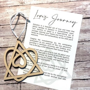 May include: Wooden ornament shaped like a heart with a Celtic knot design. The ornament is accompanied by a card with the text "Love's Journey" and a message about adoption.