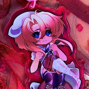 May include: A pink and white anime-style character keychain with a bloody knife. The character has blonde hair, blue eyes, and is wearing a white dress with a purple bodice. The keychain is attached to a pink metal clasp.