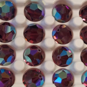 May include: A collection of faceted, round beads in a deep purple hue, arranged in a white tray. The beads have iridescent, multi-colored reflections, adding a shimmering effect. These beads are likely for jewelry making or crafting.