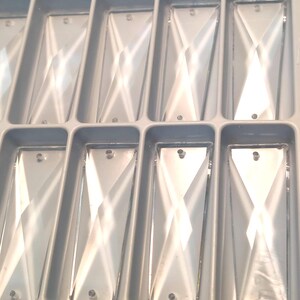 May include: A close-up shot of rectangular, clear crystal pendants with a diamond-shaped design. Each pendant has a small hole at the top for hanging. The pendants are arranged in a gray plastic tray.
