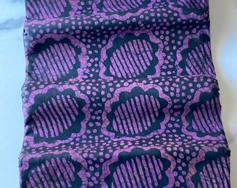 May include: Folded fabric with a repeating pattern of large, stylized floral shapes in purple and black. The design includes dotted accents between the floral motifs. The fabric is neatly folded, showcasing the pattern's repeat.